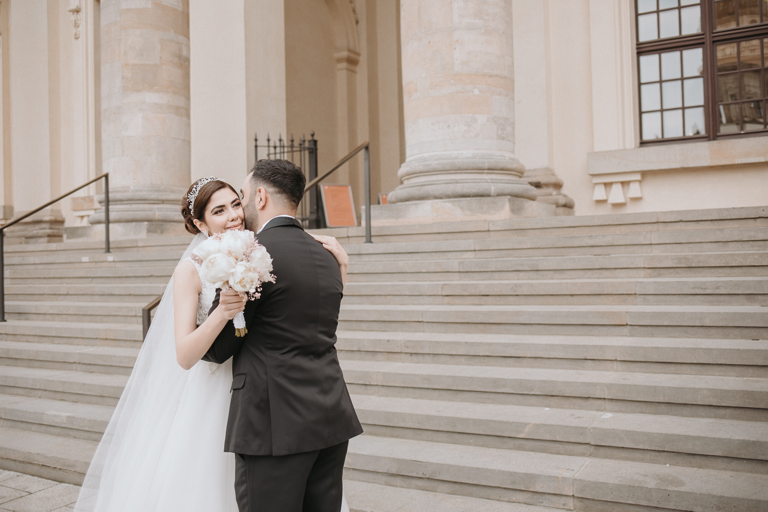 First look- Das Foto entstand wenige Sekunden nach dem Firstlook. Das Foto entstand im Rahmen des Hochzeitsshooting in Berlin vor dem dortigen Konzerthaus statt. Mann sieht das Brautpaar arm in Arm stehen. Sie stehen vor einer Sandsteintreppe. Die Braut guckt in meine Richtung, jedoch knapp an mir vorbei. Sie h